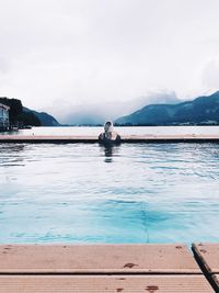 Rear view of woman swimming in infinity pool by lake against sky