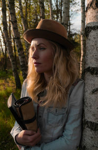 Beautiful young woman with book standing by tree trunk