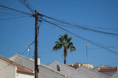 Low angle view of roof, power cables, palm and building against blue sky