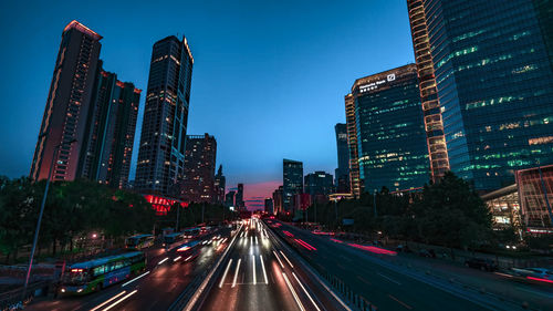 Light trails on city street by buildings against sky at night