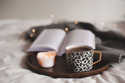 Cup of coffee with leopard print staying on wooden tray with scented burning candle and open book