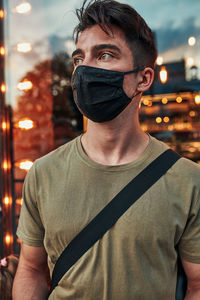 Young man standing at store front in the city center in the evening, looking away