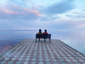 Rear view of men sitting on bench looking at sea against sky