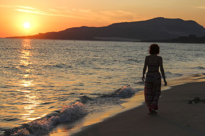 Rear view full length of woman walking on shore at beach during sunset