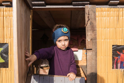 Ethnic girl in headband looking at camera with smile while standing on door on terrace of house in countryside