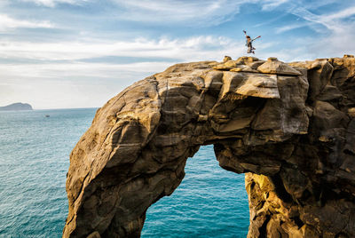 Woman jumping over rock formation at beach against sky