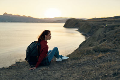 Man sitting on rock at beach against sky during sunset