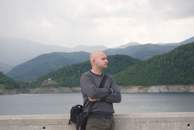 Thoughtful bald man with arms crossed standing against mountains