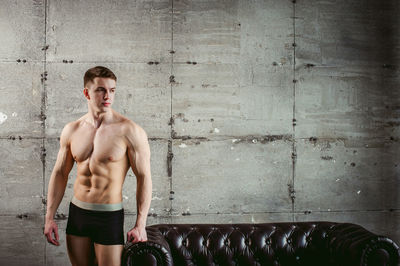 Shirtless muscular man standing by armchair against wall