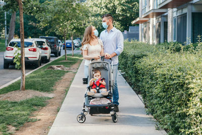 Caucasian mother and father in face masks walking with baby in stroller. family strolling together 