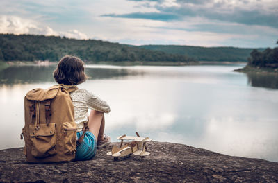 Rear view of man sitting on rock by lake against sky