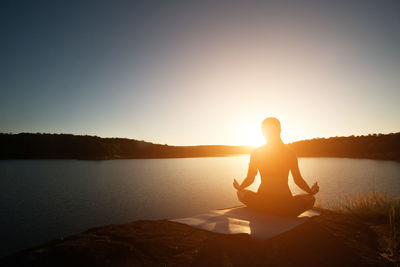 Rear view of woman meditating while sitting on rock at lakeshore during sunset