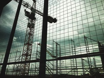 Low angle view of crane seen through fence against cloudy sky