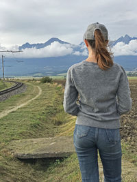 Young woman in jeans and sweater and cap looking in distance across field on mountains