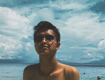 Shirtless young man wearing sunglasses at beach against cloudy sky