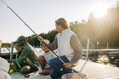 Counselors teaching fishing to kids while sitting on jetty near lake at summer camp