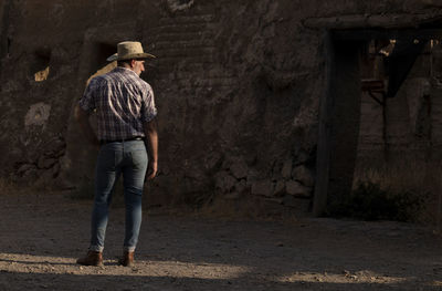 Rear view of adult man in cowboy hat and shirt against abandoned building