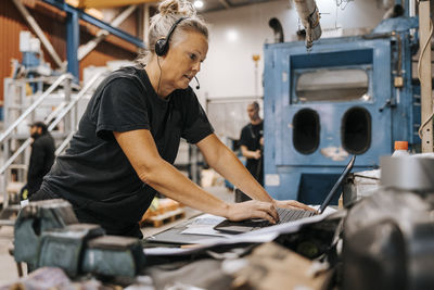 Mature female warehouse worker wearing headset while using laptop in manufacturing factory