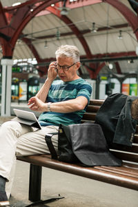 Senior commuter sitting on bench talking through smart phone while checking time at railroad station