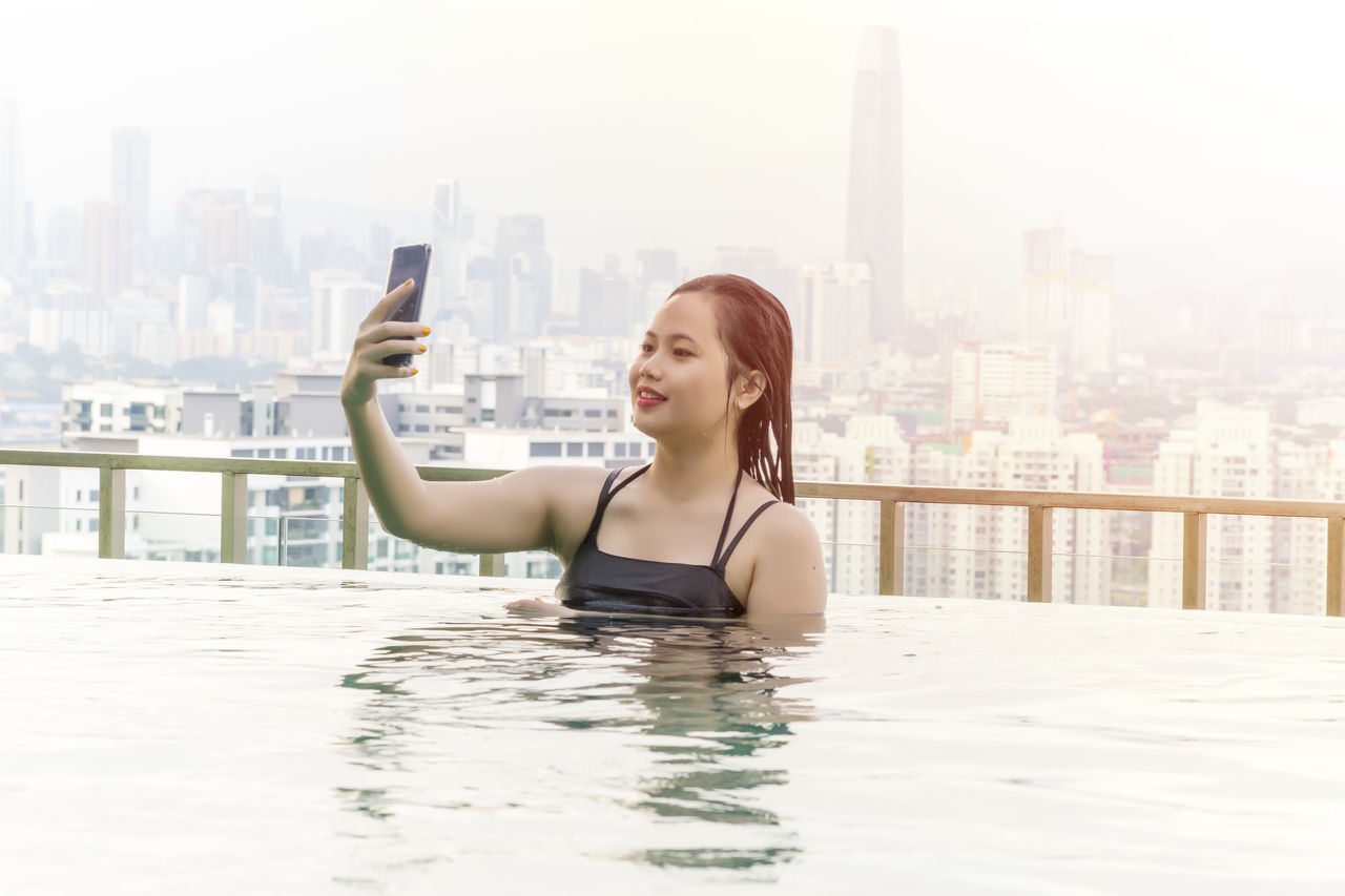 PORTRAIT OF YOUNG WOMAN USING MOBILE PHONE AT CITYSCAPE