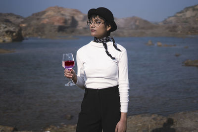 Young beautiful girl standing near a lake with a beautiful scenery in the background, holding wine.