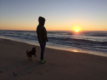 Silhouette woman by dog standing on beach against sky during sunset