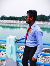 Young man looking away while standing by railing against lake