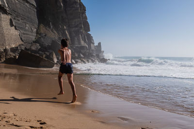 Rear view of boy in blue swim shorts running on sea shore. 