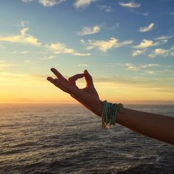 Cropped image of woman performing mudra by sea against sky during sunset