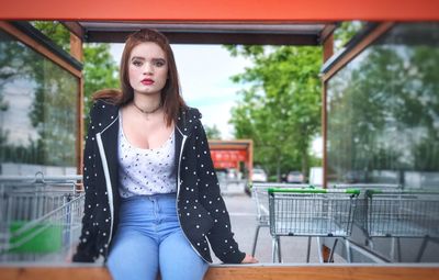 Portrait of beautiful woman sitting on retaining wall against shopping carts