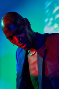 Confident bald african american male model with necklace and earring in trendy outfit standing with eyes closed at wall with glowing lights in modern studio