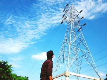 Low angle view of man looking at electricity pylon against blue sky