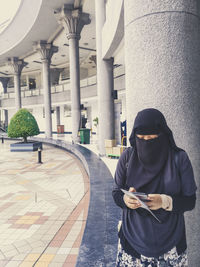 Woman wearing hijab using smart phone while standing against column