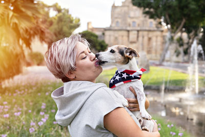 Woman holding small senior dog in park outdoor. dog is wearing bandana with an american flag