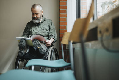 Mature man with disability reading magazine while sitting in wheelchair at waiting room