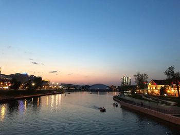 Illuminated buildings by river against sky at dusk