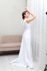 Full length portrait of bride in elegant wedding dress posing with arms raised, pensive expression 