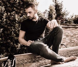 Young man looking away while sitting on tree against plants