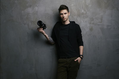 Portrait of young man holding camera while standing against wall