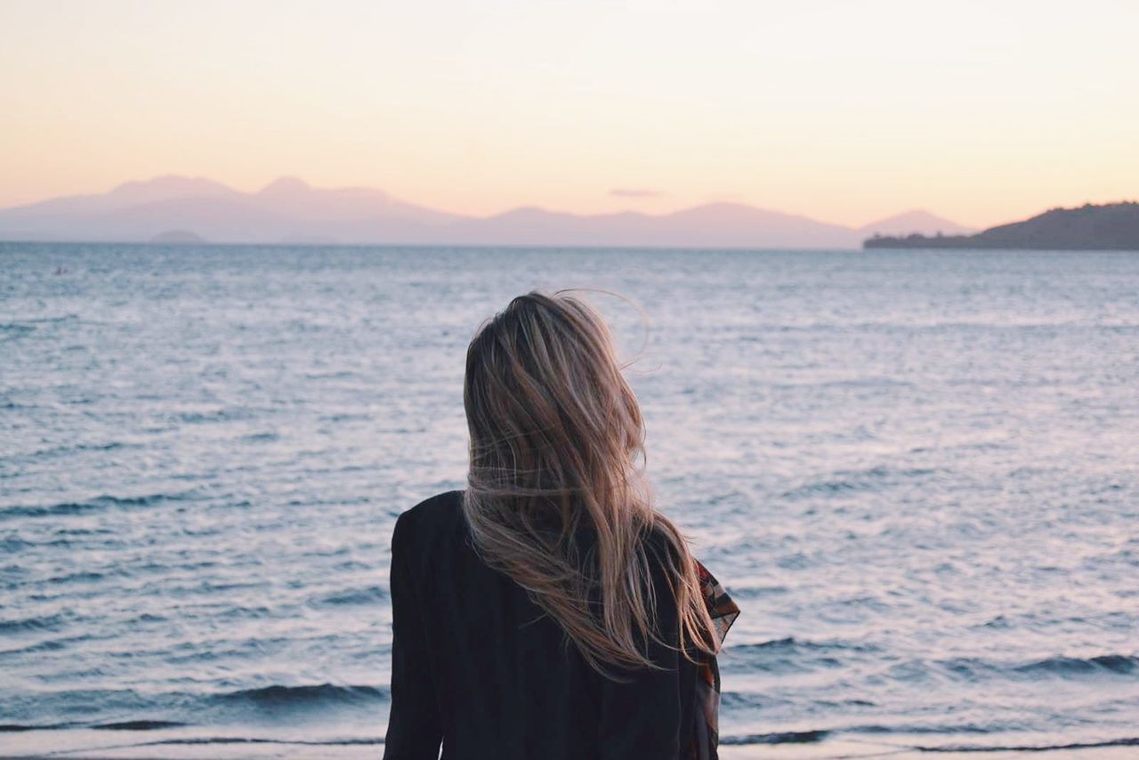 REAR VIEW OF WOMAN STANDING AT BEACH DURING SUNSET