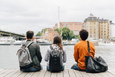 Rear view of friends with backpacks sitting on pier by river in city against sky