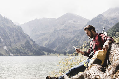 Austria, tyrol, alps, hiker relaxing on tree trunk at mountain lake checking cell phone