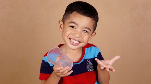 Portrait of smiling boy holding drinking glass while gesturing against wall