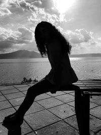 Full length of silhouette woman sitting on park bench against sea and sky during sunny day