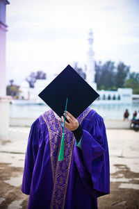 Woman wearing graduation gown covering face with mortarboard