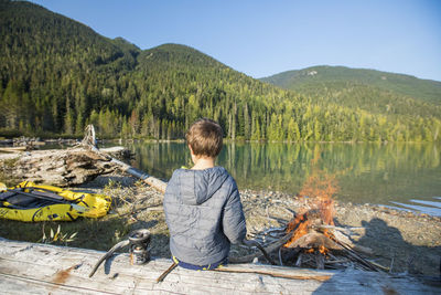 Young boy sitting on log, watching fire next to lake.
