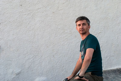 A middle-aged man sits against a white wall in summer clothes.