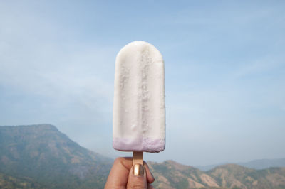 Cropped hand holding popsicle by mountain against sky