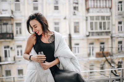 Smiling woman with wineglass wearing blanket while standing at balcony during winter