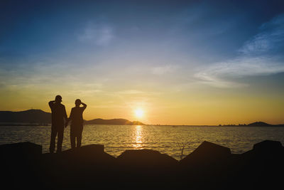 Silhouette men standing on shore against sky during sunset